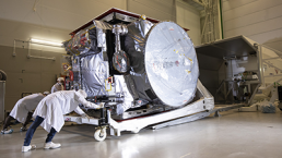 Technicians in white lab coats and caps are moving a large, cube-shaped satellite covered in protective material on a wheeled platform inside a cleanroom. The satellite, part of JUICE’s final take-off preparations before lift-off, has a circular component on one side and various equipment attached.