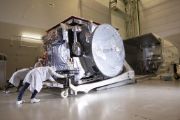 Technicians in white lab coats and hair nets work on a large, wrapped satellite within a sterile, indoor laboratory setting. As part of JUICE&rsquo;s final take-off before lift-off, one technician pushes the satellite mounted on a wheeled platform while another adjusts equipment.
