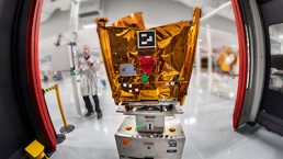 A person in a lab coat stands adjacent to a large, gold-foil-wrapped satellite or space probe inside a cleanroom. The space probe, representing constellations of opportunities, is mounted on a wheeled platform and surrounded by advanced equipment and red-framed barriers.