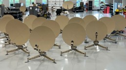 A group of large, beige satellite dishes arranged in rows inside a spacious, brightly lit industrial facility. The dishes are mounted on tripod stands with various electronic equipment visible in the background. ADSGS to provide Ranger 1200 Satellite Terminals to U.S. Army amid these advanced setups.
