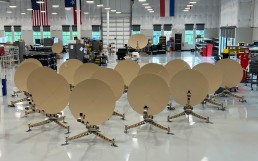 A room with numerous identical gold-colored satellite dishes arranged in rows, each mounted on tripod stands. The space appears to be a manufacturing or assembly facility, with workbenches, equipment, and windows in the background, alongside hanging flags. ADSGS to Provide Ranger 1200 Satellite Terminals to U.S. Army.