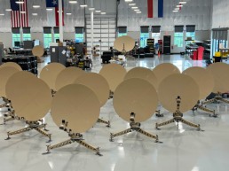A room with numerous identical gold-colored satellite dishes arranged in rows, each mounted on tripod stands. The space appears to be a manufacturing or assembly facility, with workbenches, equipment, and windows in the background, alongside hanging flags. ADSGS to Provide Ranger 1200 Satellite Terminals to U.S. Army.