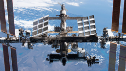 The image shows the International Space Station (ISS) orbiting above Earth, with its large solar panels and various modules, including Bartolomeo – The Easy Way of Bringing Payloads to the ISS. Below, Earth's surface features distinguishable landmasses and bodies of water under a partly cloudy sky.