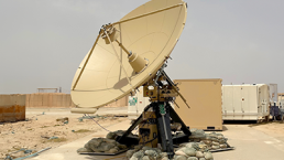 A large beige satellite dish, part of the ADSGS providing RCTs (Rapid Capability Terminal) to the U.S. Space Force, is mounted on a black base surrounded by sandbags. It sits outdoors on a concrete pad in a desert-like environment, with storage containers and structures visible under a hazy sky.