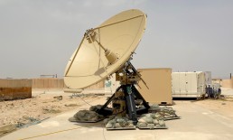 A large satellite dish, part of the ADSGS to provide RCT&rsquo;s to the U.S. Space Force, stands outdoors on a concrete surface surrounded by sandbags for stability. The background reveals some shipping containers and a desert-like landscape under a clear sky.