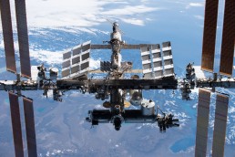 A detailed view of the International Space Station with Earth in the background. The station's large solar panels and various modules, including Bartolomeo &ndash; The Easy Way of Bringing Payloads to the ISS, are clearly visible. The Earth below shows large landmasses and bodies of water, partly covered by clouds.
