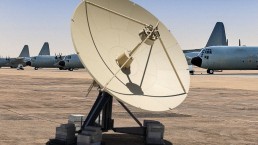 A large satellite dish on the ground with several military aircraft, likely C-130 Hercules planes, lined up in the background. An Airbus RCT 4.0M is situated nearby. The sky is clear and the area appears to be a military airbase.
