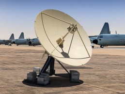 A large satellite dish on the ground with several military aircraft, likely C-130 Hercules planes, lined up in the background. An Airbus RCT 4.0M is situated nearby. The sky is clear and the area appears to be a military airbase.