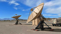 Two large satellite dishes are mounted on metal frames in a desert landscape. The sky above is blue with scattered clouds. A portable generator is located next to one of the satellite dishes. Hills can be seen in the background, showcasing where the U.S. Marines choose Airbus RCTs for their operations.
