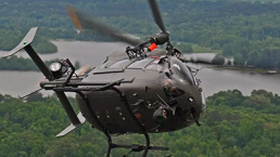 A military helicopter, part of the U.S. Army's recent Airbus contract for helicopter modernization, flies above a dense forested area and a large body of water. Painted in dark colors, its rotors blur with motion over the landscape below, featuring a mix of green trees and winding waterways.