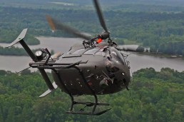 A U.S. Army helicopter flies in the sky above a lush green forested area with a river in the background. The helicopter, part of the recent Airbus contract for modernization, features a dark exterior and various attachments, captured in motion as it navigates through the air.