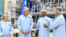         Four engineers wearing blue lab coats and hairnets are having a discussion in a laboratory environment with high-tech equipment in the background. The laboratory appears to be related to aerospace or advanced technology, possibly as Artemis II astronauts discover a key component of the Orion spacecraft.
