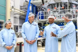 Four individuals, wearing blue lab coats, hair nets, and white protective coverings, stand inside a high-tech manufacturing facility. One person is gesturing while speaking with the others as the Artemis II astronauts discover a key component of the Orion spacecraft. Equipment with 