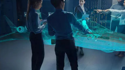 A group of five people in a futuristic control room observe and interact with a large, blue holographic model of an advanced aircraft, part of the Future Combat Air System (FCAS). The background features illuminated computer panels and screens that seamlessly integrate with the Internet of Military Things.