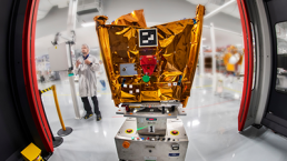 A scientist in a lab coat stands in a cleanroom beside a spacecraft covered in gold foil, mounted on a platform with electronic equipment. The room, featuring red-framed glass doors and various scientific instruments, buzzes with news: Airbus announces the purchase of Eutelsat OneWeb’s stake in satellite constellation manufacturing joint venture.