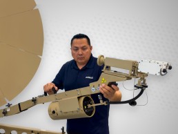 A person wearing a navy blue Airbus polo shirt is handling a large, beige mechanical apparatus from the Family of Terminals against a light grey background. The apparatus appears complex with various components and wires. A partial view of a circular object with triangular segments is visible on the left.