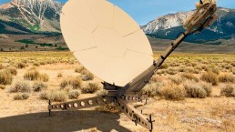 A tan satellite dish with an intricate support structure is situated in a vast, dry grassy field with mountains in the background under a clear blue sky. The dish, part of Airbus DS Government Solutions, is pointed slightly upwards and to the right, with its metallic support casting a shadow on the ground.
