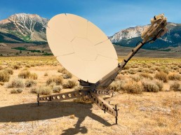 A tan satellite dish with an intricate support structure is situated in a vast, dry grassy field with mountains in the background under a clear blue sky. The dish, part of Airbus DS Government Solutions, is pointed slightly upwards and to the right, with its metallic support casting a shadow on the ground.
