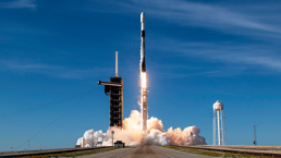 A rocket carrying the EUTELSAT 36D Airbus-built satellite is launching into a clear blue sky from a launch pad. Smoke and steam billow from the base of the rocket, and a water tower is visible to the right of the pad. The scene is captured in bright daylight.